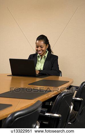 Stock Image - Businesswoman sitting at conference table smiling and typing on laptop computer.. Fotosearch