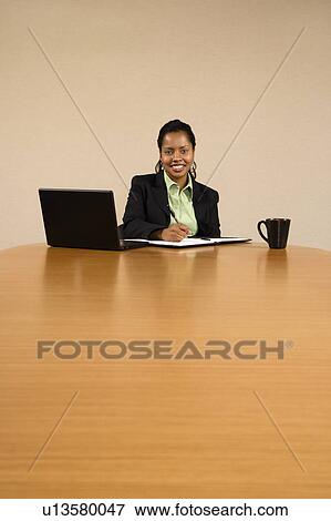 Businesswoman sitting at conference table with laptop computer and coffee cup smiling and writing in book View Large Photo Image Stock Photo - Businesswoman sitting at conference table with laptop computer and coffee cup smiling and writing in book. Fotosearch