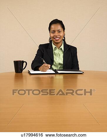 Businesswoman sitting at conference table smiling and writing in notebook View Large Photo Image Stock Photograph - Businesswoman sitting at conference table smiling and writing in notebook. Fotosearch