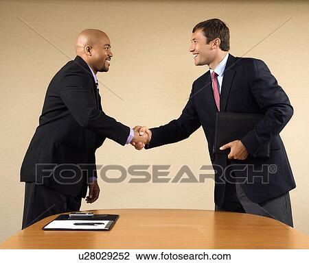 Stock Image - Two businessmen in suits shaking hands and smiling. Fotosearch