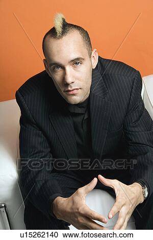 Stock Image - Caucasian man in suit with mohawk sitting in chair against orange background.. Fotosearch