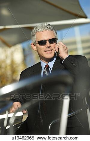 Stock Photography - Prime adult Caucasian man in suit sitting at ouside patio table wearing sunglasses and talking on cellphone smiling.. Fotosearch