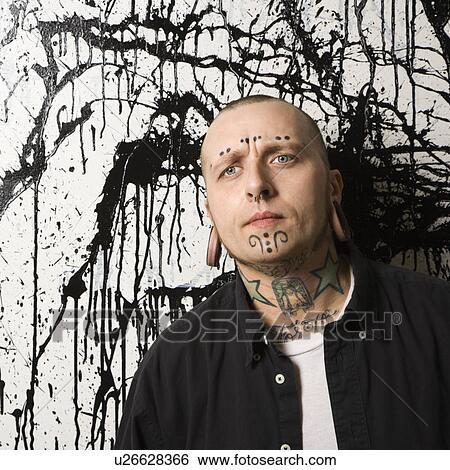 Stock Photograph - Tattooed and pierced man standing against paint splattered background.. Fotosearch