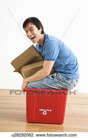 Asian young man trying to sit in recycling bin with cardboard. View Large Photo Image Stock Image - Asian young man trying to sit in recycling bin with cardboard.. Fotosearch