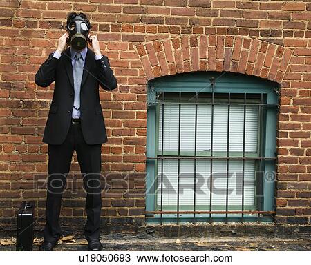 Stock Image - Businessman standing next to brick wall wearing gas mask.. Fotosearch