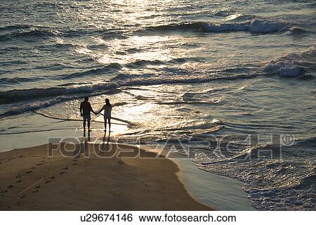 Stock Photograph - Aerial view of couple holding hands on beach in Bald Head Island, North Carolina at sunset. Fotosearch