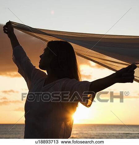 Attractive Caucasian mid-adult woman holding up fabric in breeze silhouetted by sunset beside ocean. View Large Photo Image Stock Image - Attractive Caucasian mid-adult woman holding up fabric in breeze silhouetted by sunset beside ocean.. Fotosearch