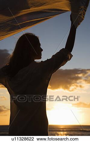 Attractive mid-adult woman holding up fabric in breeze silhouetted by sunset beside ocean View Large Photo Image Stock Image - Attractive mid-adult woman holding up fabric in breeze silhouetted by sunset beside ocean. Fotosearch