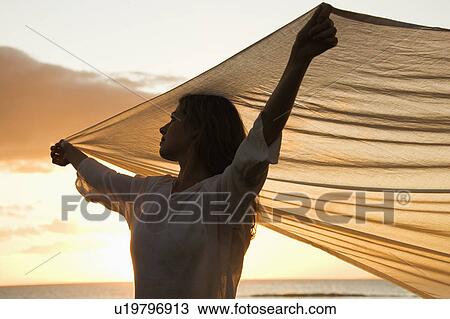 Stock Image - Attractive woman holding up fabric in breeze silhouetted by sunset beside ocean. Fotosearch