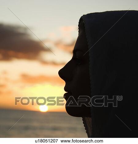 Stock Photo - Caucasian mid-adult woman with scarf over head profiled against sunset over ocean.. Fotosearch