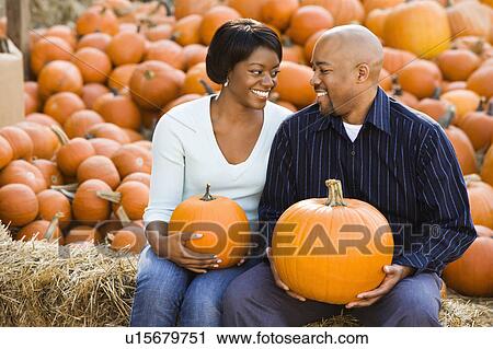 Stock Image - Couple sitting on hay bales holding pumpkins. Fotosearch