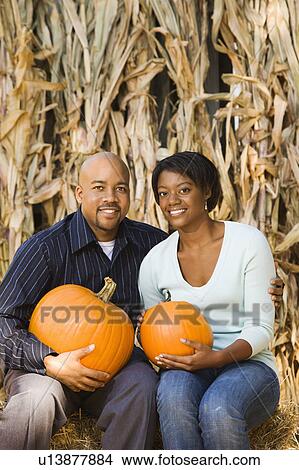 Picture - Happy smiling couple sitting on hay bales and holding pumpkins at outdoor market.. Fotosearch