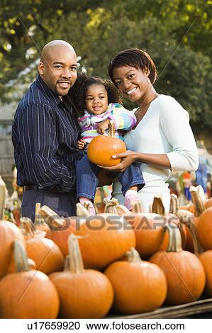 Stock Image - Parents and daughter picking out pumpkin and smiling at outdoor market. Fotosearch