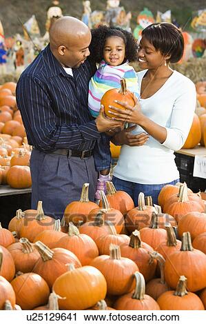Stock Image - Parents and daughter picking out pumpkin and smiling at outdoor market.. Fotosearch