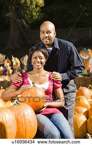 Picture - Portrait of happy smiling couple sitting in pumpkins at outdoor market.. Fotosearch