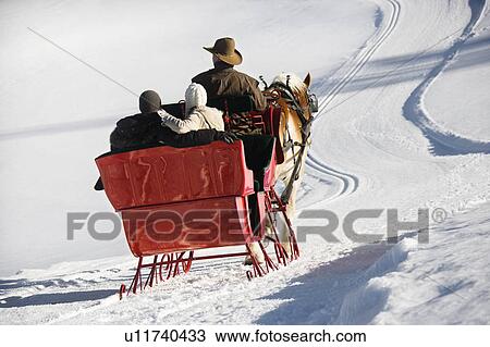 Stock Image - Rear view of Caucasian man and young couple riding in horse-drawn sleigh in snow.. Fotosearch