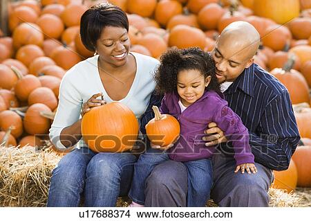 Picture - Smiling family sitting on hay bales and holding pumpkins. Fotosearch
