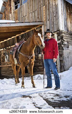 Stock Photo - Young Caucasian man in western attire holding his horse with stable in background.. Fotosearch