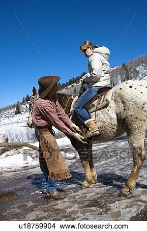 Picture - Caucasian male wrangler helping young adult Caucasian female on horseback.. Fotosearch