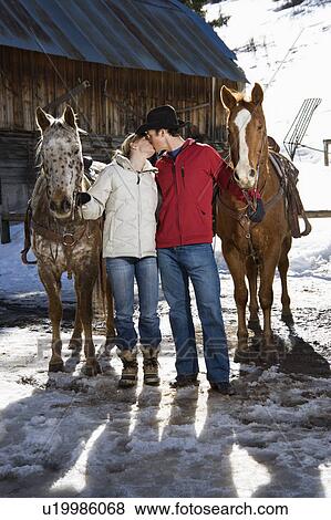 Stock Photo - Couple kissing and holding horses with stable in background.. Fotosearch