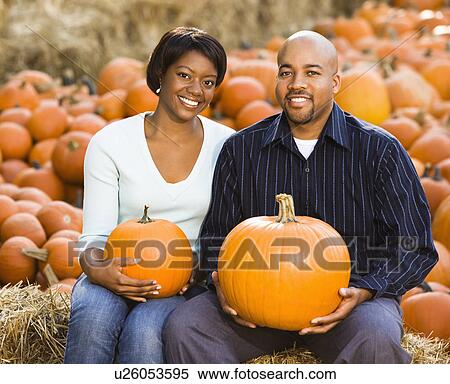 Stock Photography - Happy smiling couple sitting on hay bales and holding pumpkins at outdoor market.. Fotosearch