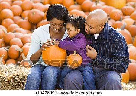 Stock Image - Happy smiling family sitting on hay bales and holding pumpkins at outdoor market. Fotosearch