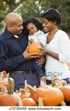 Stock Photo - Parents and daughter picking out pumpkin and smiling at outdoor market. Fotosearch