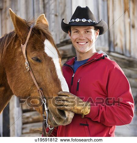 Stock Image - Young Caucasian man in cowboy hat petting his horse.. Fotosearch