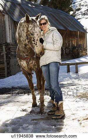 Young woman petting horse with stable in background in winter View Large Photo Image Stock Photo - Young woman petting horse with stable in background in winter. Fotosearch