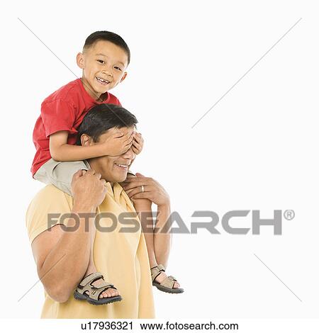 Stock Image - Asian boy sitting on father's shoulders with hands over his eyes in front of white background.. Fotosearch