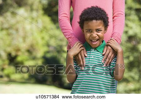 Boy in park making funny expression with mother's hands on shoulders. View Large Photo Image Picture - Boy in park making funny expression with mother's hands on shoulders.. Fotosearch