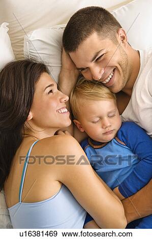 Stock Photo - Caucasian mid adult parents cuddling with toddler son sleeping in bed.. Fotosearch
