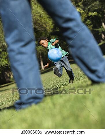 Stock Photography - Son running and kicking ball towards father in park. Fotosearch