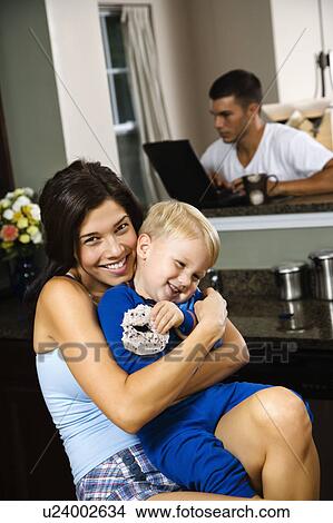 Picture - Woman hugging toddler son with father on laptop in background. Fotosearch
