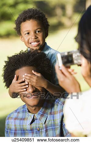 Stock Image - Woman taking digital photo of husband and son in park.. Fotosearch