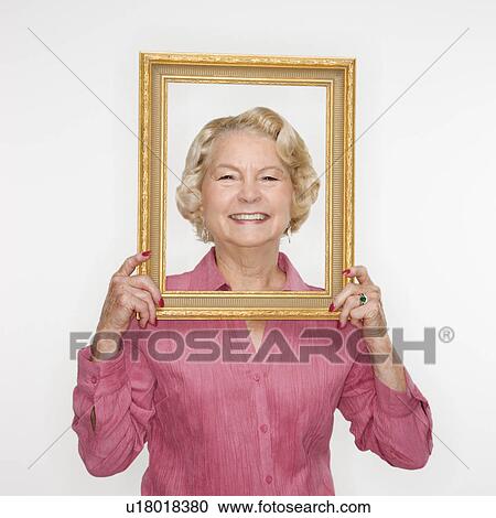 Stock Image - Caucasian senior woman holding picture frame over face.. Fotosearch