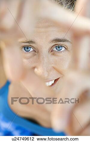 Stock Image - Close-up of Caucasian mid-adult woman smiling and looking at viewer through framed fingers.. Fotosearch