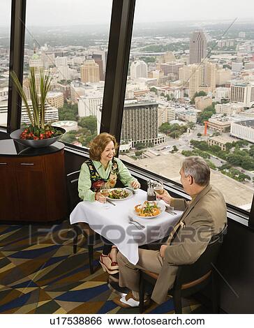 Stock Photograph - Mature couple dining in fancy restaurant by window with rooftop view of urban landscape. Fotosearch