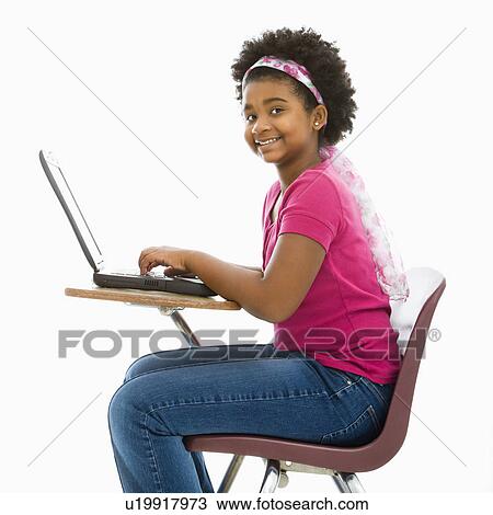 Stock Image - African American girl sitting in school desk typing on laptop computer smiling at viewer.. Fotosearch