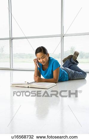 Stock Image - Asian preteen girl lying on floor doing homework while talking on cell phone.. Fotosearch
