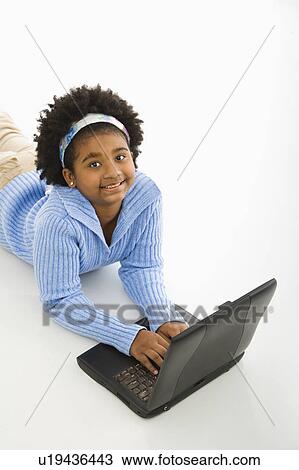 Stock Image - High angle view of girl laying on floor using laptop and looking up at viewer. Fotosearch
