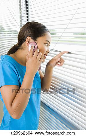 Picture - Side view of girl looking through blinds out window while talking on cell phone. Fotosearch