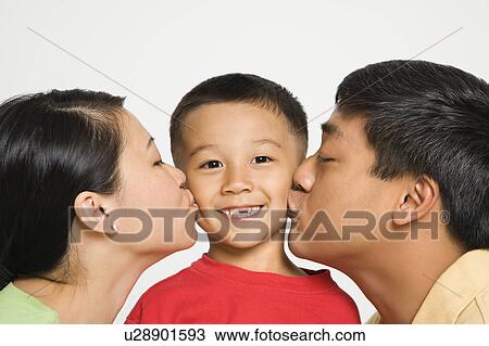 Stock Image - Asian mother and father kissing opposite cheeks of smiling son in front of white background.. Fotosearch