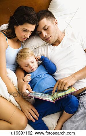 Stock Image - Caucasian parents and toddler son reading book in bed.. Fotosearch