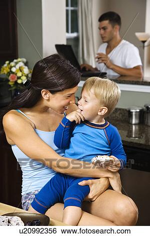Stock Photograph - Caucasian woman with toddler son in kitchen with father on laptop in background.. Fotosearch