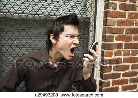 Stock Photo - Angry young Asian man next to brick wall and window yelling at cell phone.. Fotosearch