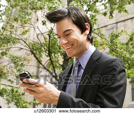 Stock Image - Asian business man standing looking at cell phone messages laughing.. Fotosearch