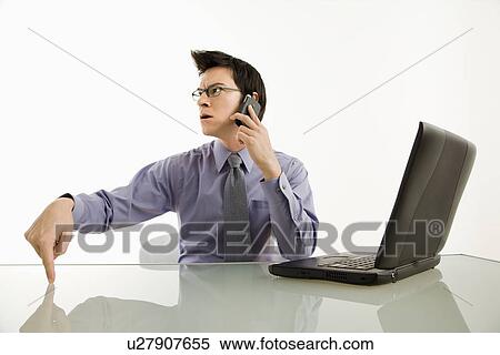 Stock Photography - Businessman sitting at desk on cellphone with angry expression. Fotosearch