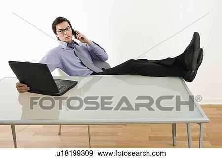 Businessman sitting at desk talking on cellphone and using laptop View Large Photo Image Stock Photo - Businessman sitting at desk talking on cellphone and using laptop. Fotosearch