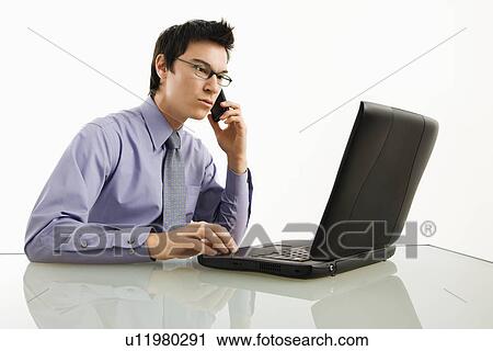 Businessman sitting at desk working on laptop talking on cellphone View Large Photo Image Stock Image - Businessman sitting at desk working on laptop talking on cellphone. Fotosearch